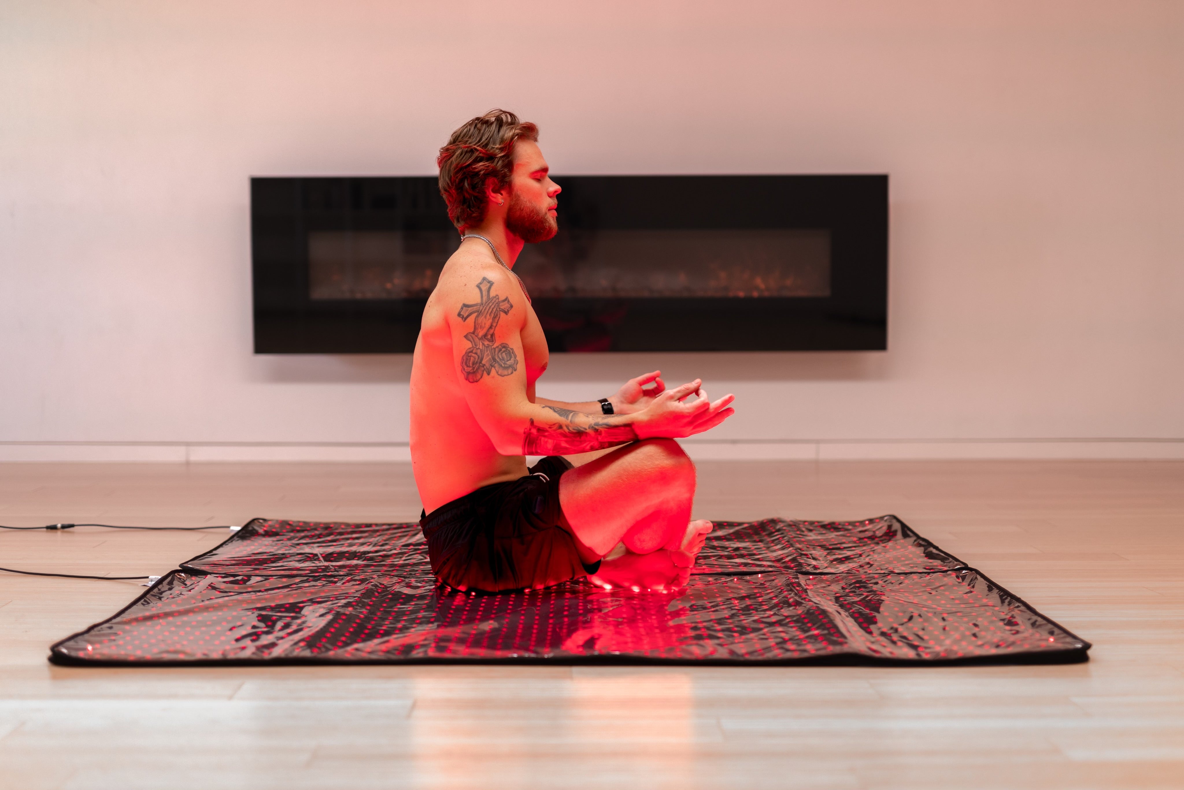 Man sits cross-legged meditating on the open BioLight Cocoon pod glowing red on a wood floor